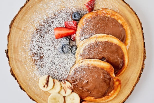 three american style pancakes on wooden board, topped with Nutella and fruit