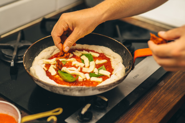 Hands adding ingredients to pizza in a frying pan