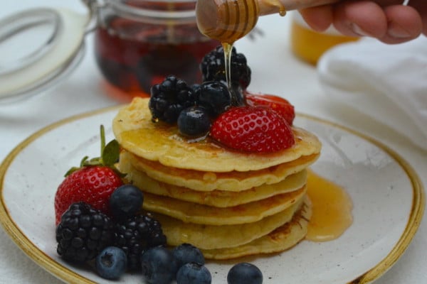 stacks of pancakes on a plate topped with berries. someone is pouring honey on top