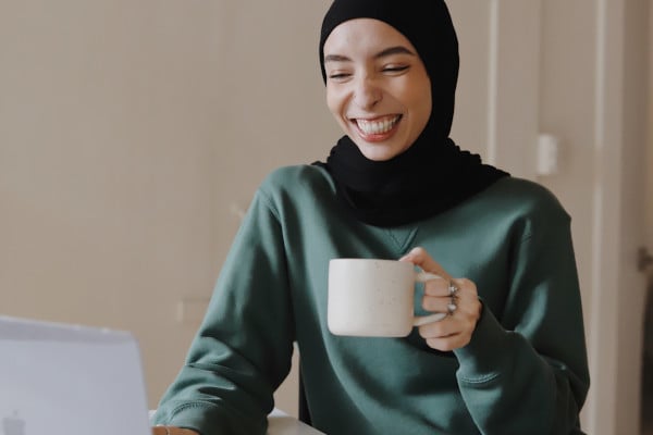 Woman in hijab smiling at laptop with mug of coffee